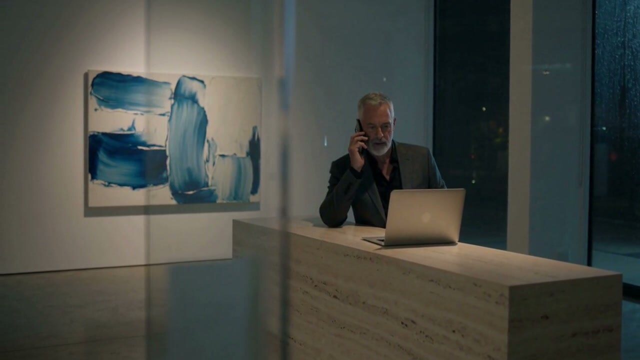 A gallery owner on the phone at his desk at night, a blue painting on the wall behind him